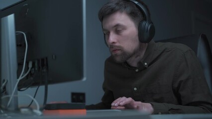 Application developer in wireless headphones working. sitting at computer workplace in home office. IT guy focused and tiredly looking at monitor while programming. Overtime, deadlines are on fire.