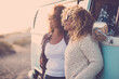 © simona - Two adult young woman enjoying time and leisure together against a classic blue van in travel vacation activity admiring the sunset. Friendship and female people traveler. Freedom emotion vanlife.