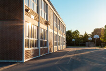 The Exterior Of The School Building And School Yard With A Basketball Court On A Sunny Evening. The Sun Ireflecting In The Windows