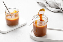 Liquid Caramel On Spoon On Glass Jar. Two Glass Jars With Caramel Sauce. Close Up. Grey Napkin And White Background