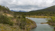 Landschaftsidyll, ein Fluss schlängelt sich durch die grüne Graslandschaft Patagoniens, am Horizont ragen hohe, schneebedeckte Berge empor, welche in der weißen Wolkendecke verschwinden 