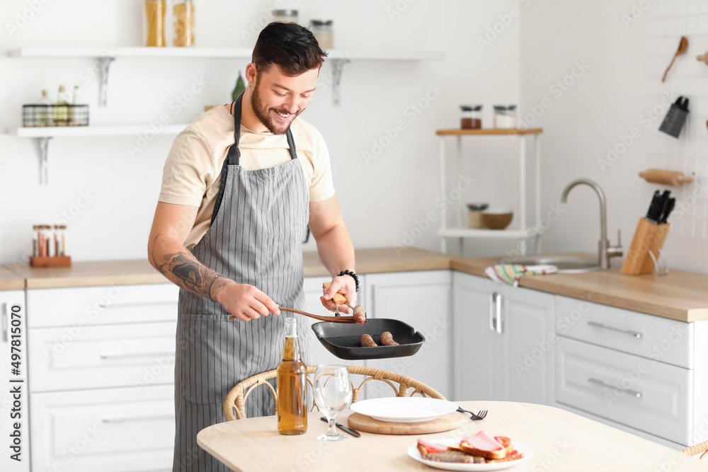 Young bearded man putting fried sausages onto plate in kitchen