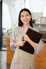 Pretty Asian barista in apron holding a clipboard and standing in front of a coffee shop counter. Startup concept of business owner.