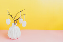 Happy Easter Holiday, Coming Spring Concept. Tree Branches, Flower Twigs In White Vase Or Jar With Hanging Eggs. Yellow And Pink Table Background With Copy Space For Ad Or Text. Easter Home Decoration