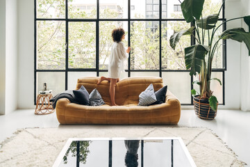 Young african american woman standing on couch at home looking out the window.