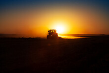 Tractor In Sunset, Farmer Ploughs A Field With Tractor In Sunset.  The Tractor Is Backlit By The Setting Sun.  Day Is Turning Night In The Field. Beautiful Agriculture Concept Idea Photo.