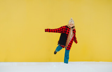 Wall Mural - Funny teen girl in casual clothes having fun outdoors in winter, standing on one leg in the snow on a background of yellow wall.