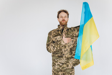 Ukrainian patriot soldier in military uniform holds a hand on a heart with a yellow and blue flag on a white background