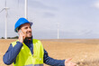 © Dani D.G - Adult man dressed in work clothes, safety helmet and reflective vest, smiling, talking on his mobile phone with his co-worker at a wind farm. Security technician in a field with wind generators.