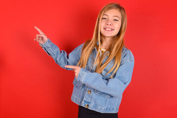 Wall Mural - blonde little kid girl wearing denim jacket over red background indicating finger empty space showing best low prices, looking at the camera