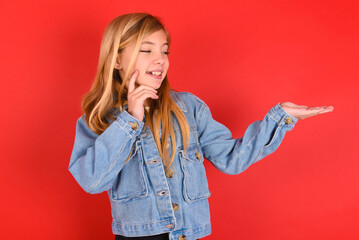 Wall Mural - Funny blonde little kid girl wearing denim jacket over red background holding open palm new product. I wanna buy it!