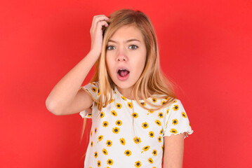 Wall Mural - Embarrassed blonde little kid girl wearing daisy t-shirt red over background with shocked expression, expresses great amazement, Puzzled model poses indoor