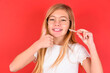© Roquillo - blonde little kid girl wearing denim jacket over red background holding an invisible braces aligner and rising thumb up, recommending this new treatment. Dental healthcare concept.