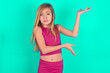 blonde little kid girl wearing pink sport clothes over green background pointing aside with both hands showing something strange and saying: I don't know what is this. Advertisement concept.