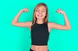 Strong powerful blonde little kid girl wearing black sport clothes over green background toothy smile, raises arms and shows biceps. Look at my muscles!