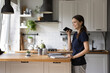 © fizkes - Young woman standing in domestic kitchen holding smart phone leaves voice message, share voicemail to friend, having pleasant conversation use speakerphone, easy convenient modern tech usage concept