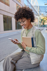 Wall Mural - Happy curly haired woman uses mobile application for browsing media content reads receieved message holds textbooks wears jumper vest and trousers sits outdoors during sunny day. Technology concept