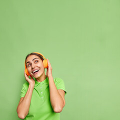 Wall Mural - Vertical shot of positive young woman keeps hands on stereo headphones looks above dressed in casual t shirt isolated over vivid green background blank copy space for your promotional content