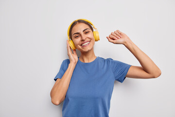 Wall Mural - Waist up shot of positive carefree European woman dances and catches every bit of music wears casual blue t shirt listens favorite song via wireless headphones isolated over white background.