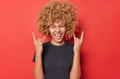 © wayhome.studio  - Positive young curly woman shows horn gesture enjoys rock music wears black casual t shirt isolated over vivid red background. Overjoyed curly haired female model demonstrates heavy metal sign