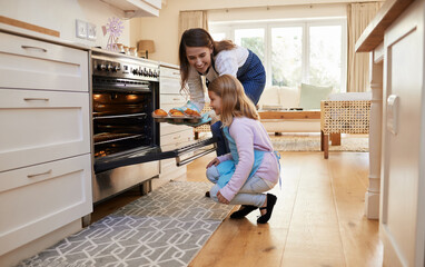 Poster - Be careful its very hot. Shot of a mother and daughter inserting a tray of cupcakes into the oven.