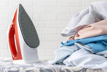 Close-up Of A Stack Of Washed, Rumpled Clothes And A Hot Steam Iron On An Ironing Board. The Concept Of Household Chores, Daily Routine, Clothes Care.