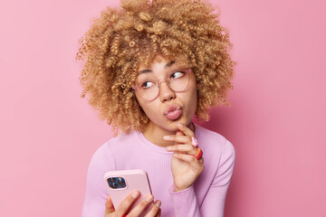 Wall Mural - Beautiful dreamy woman keeps lips folded looks away wears transparent glasses and casual jumper uses smartphone waits for call isolated over pink background. Lovely young female with gadget.