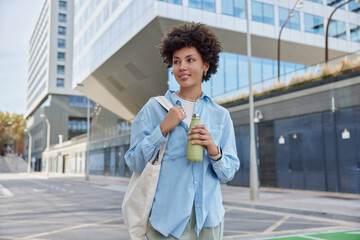 Wall Mural - Young beautiful curly haired woman wears denim shirt carries fabric bag holds bottle and drinks water looks happily away poses in city centre returns from work. Urban street lifestyle concept