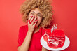 Happy overjoyed young woman covers mouth with hand eats delicious creamy cake has fun laughs positively wears festive dress isolated over vivid red background. Crazy female enjoys tasty dessert