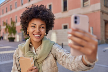 Wall Mural - Happy curly haired woman makes selfie via smartphone smiles broadly holds notebooks wears shirt and jumper tied over neck smiles broadly strolls outdoors has cheerful expression. Technology user
