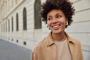 Wall Mural - outdoor shot of happy woman with curly hair dressed in brown jacket smiles broadly has good mood str