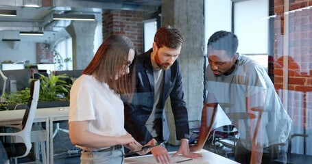 Wall Mural - Multiethnic team of IT startup having conversation in coworking space and working with documents, charts and graphics. Coworkers collaborating in office. Young workers brainstorming behind the glass.