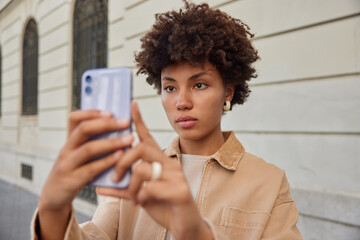 Wall Mural - outdoor shot of attractive woman with curly hair uses front mobile camera for clicking selfie images