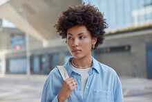 Outdoor Shot Of Beautiful Woman With Dark Curly Hair Wears Blue Casual Shirt Carries Bag Looks Away Strolls At Street Against Blurred Background During Daytime. People And Lifestyle Concept.