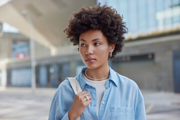 Wall Mural - Outdoor shot of beautiful woman with dark curly hair wears blue casual shirt carries bag looks away strolls at street against blurred background during daytime. People and lifestyle concept.