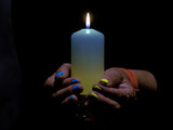 Fototapeta  - Płonąca świeca trzymana w kobiecych dłoniach. Świeca ma odcień niebieski i żółty. Na takie same kolory pomalowane są paznokcie. Jest to symbol solidarności z ofiarami wojny w Ukrainie,
