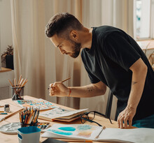 A Creative Person Is Standing Over A Desktop With A Brush In His Hand. A Young Artist Makes A Sketch With A Brush In The Studio. Creative Studio, Lifestyle, The Process Of Creating A Work Of Art