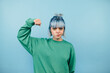 © bodnarphoto - Serious teen girl in green sweatshirt stands on blue background and shows biceps to camera.