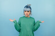 © bodnarphoto - Confused teen girl with blue hair in a green sweatshirt stands on a blue background and bewilderedly spreads his arms.