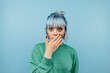 © bodnarphoto - Close portrait of a shocked girl with blue hair stands on a blue background and looks at the camera with a surprised face covering his mouth with his hand in shock.