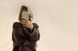 © Julia - Portrait of a beautiful blonde woman in a brown sweater covering her face with a smartphone, beige background, day