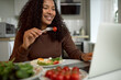 © Anatoliy Karlyuk - Happy joyful young African American woman with long curly hair in brown sweater sitting in front of opened laptop, laughing while holding fork with cherry tomato, watching video during breakfast