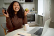 © Anatoliy Karlyuk - Cheerful joyful African girl of 20s having emotional happy face expression, smiling widely with closed eyes, holding phone in one hand and apple in another, feeling lucky, laptop and cup on table