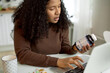 © Anatoliy Karlyuk - Pretty charming African American female nutritionist sitting at kitchen table working from home holding bottle of dietary supplements, writing article on laptop, bunch of pills lying on table