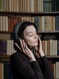 Fototapeta Zwierzęta - Woman in headphones listening to audiobooks on background of library shelves with paper books