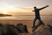 Carefree Man Jumping From Sand Dune With Arms Outstretched At Beach