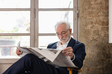Senior Man Reading Newspaper Sitting In Front Of Window At Home
