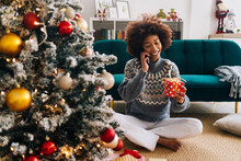 Happy Woman With Christmas Present Talking On Mobile Phone In Living Room