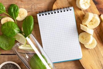 Wall Mural - Notebook with ingredients for cooking detox smoothie. Glass with spinach, banana, lime and chia seeds on wooden table top view. Healthy food concept
