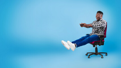 Wall Mural - Excited young black guy with joystick sitting on chair, playing video game on blue background, banner with copy space